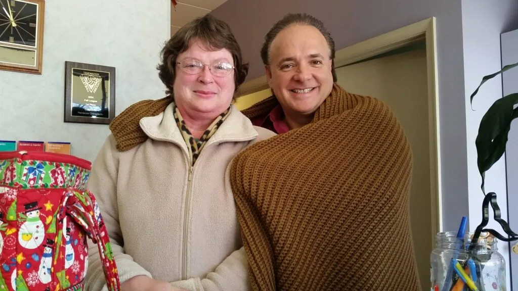 Two people posing together indoors, woman on left wearing cream colored fleece jacket with glasses, man on right wearing brown sweater, smiling at camera
