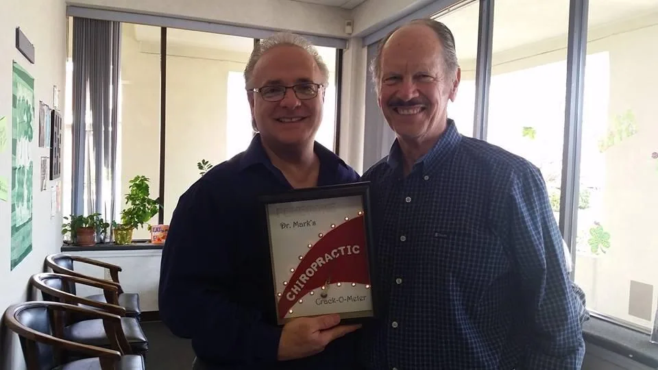 Two men standing together in an office holding a framed chiropractic sign