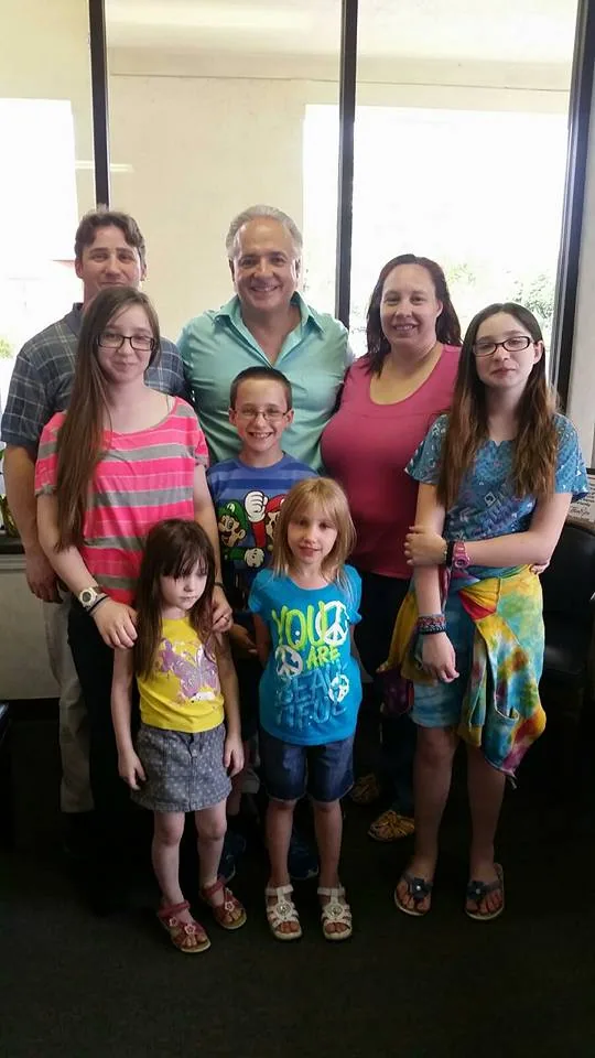 Family group of eight people posing together indoors with large windows behind them