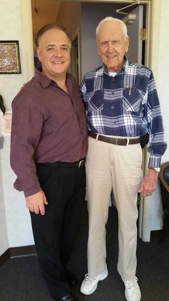 Two men standing together indoors, one wearing a maroon polo shirt and the other wearing a blue and white plaid shirt