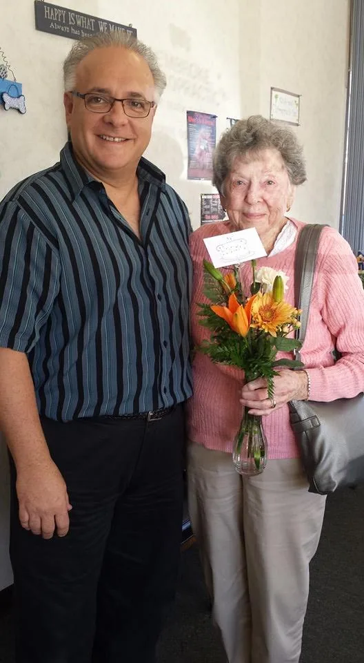 Man in striped shirt standing next to older woman in pink cardigan holding bouquet of orange and yellow flowers