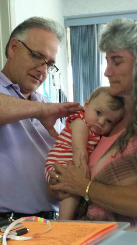 Man in glasses and light blue shirt examines young child's arm while woman holds child, indoors with window blinds visible behind them.